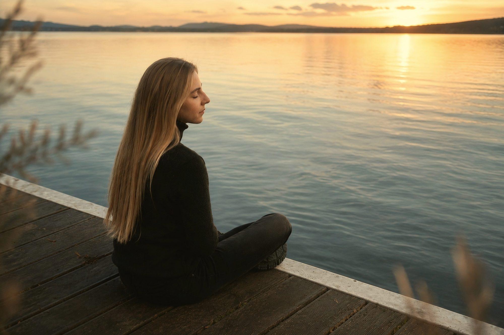 A woman sitting quietly on a wooden dock beside still water at dusk.