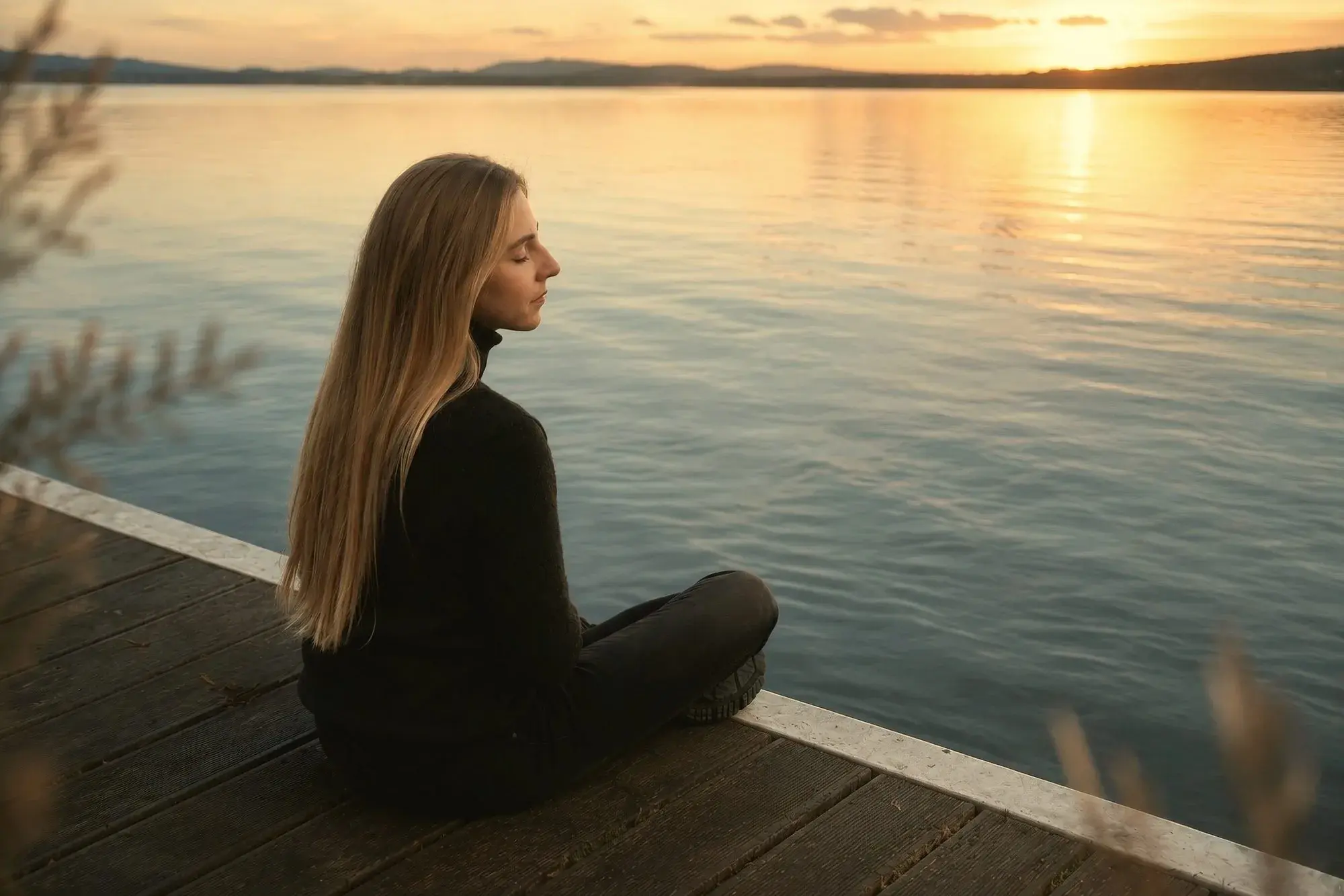 A woman sitting quietly on a wooden dock beside still water at dusk.